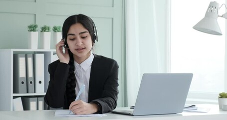 Indian businesswoman, dressed in a black blazer and white shirt, sits in her office wearing headphones. As she listens to music, she seamlessly juggles multiple tasks, including working on paperwork.