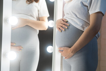 Unrecognizable pregnant woman with big belly in front of mirror, standing in room.