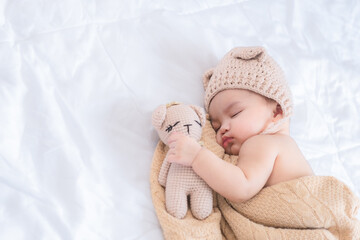 Cute baby in brown knitted hat bear style with blanket sleeping on white bed hugging bear door with peacfully and comfortably emotion.