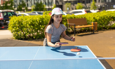 Young teenager girl playing ping pong. She holds a ball and a racket in her hands. Playing table tennis outdoors in the yard
