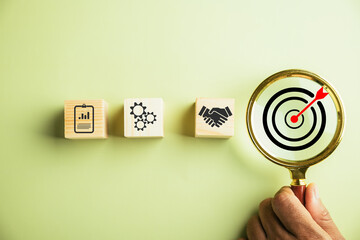 The photograph features a businessman using a magnifying glass to examine the target goal icon,...