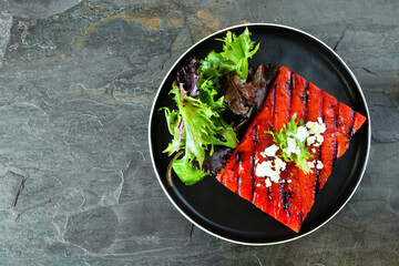 Grilled watermelon steak with salad. Above view on a dark slate background. Healthy eating, plant based meat substitute concept.