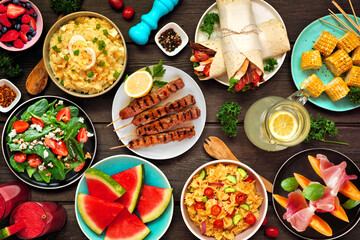 Summer food table scene over a dark wood background. Selection of refreshing salads, fruit, wraps and BBQ grilled skewers. Above view.
