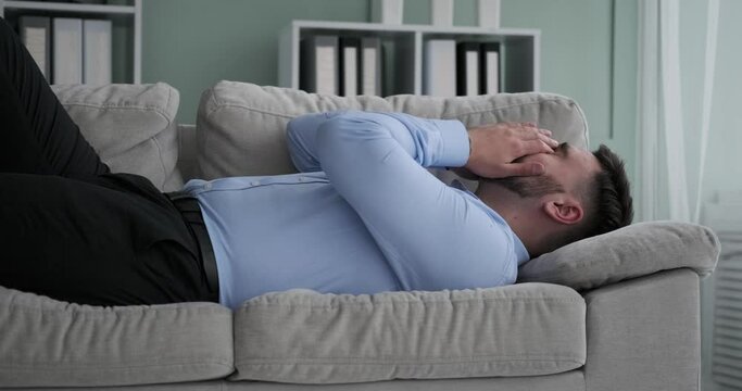 After a hard day at work, a weary Caucasian office worker returns home and plops down on the couch fully clothed. He quickly falls asleep, exhausted from the day's stresses and strains.
