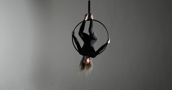 A confident Caucasian dancer captivates on the aerial hoop, defying gravity with her acrobatic skills. Against a dark backdrop, illuminated by contrasting lights.