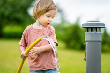 Cute toddler boy watering flower beds in the garden at summer day. Child using garden hose to water...