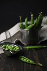 young green peas in a tin mug and nearby lie peas and pods in a sieve on a dark wooden background. for screensavers, labels, postcards, flyers, etc.