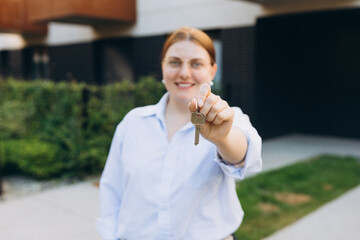 Attractive young woman showing key while standing outdoor against new house. Real estate agent holding key on street background