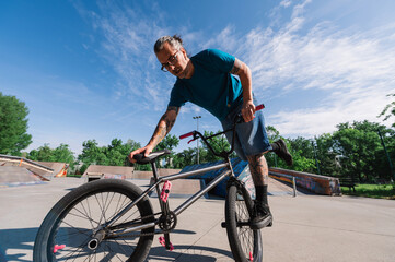 A middle-aged tattooed active man is balancing on on foot on his bmx in a skate park.