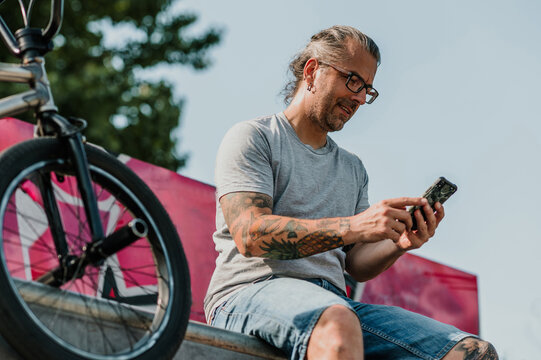 A middle-aged tattooed contemporary man is sitting in a skate park and following feed on social media.