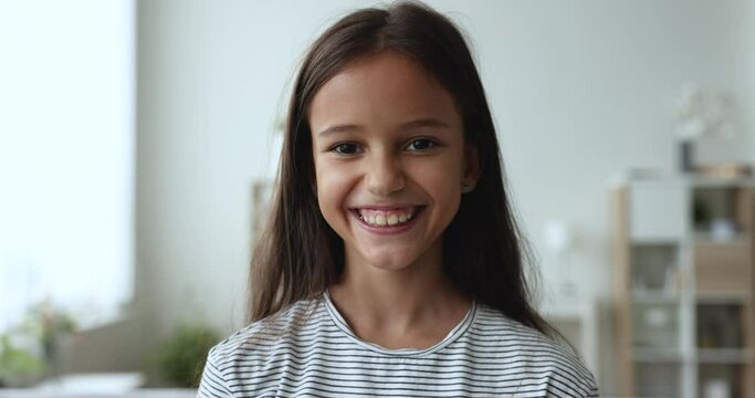 Close up head shot of cheerful beautiful little pre-teen 8s-10s girl standing in living room smile look pose for camera at home. Portrait of happy young Z or Alpha generation kid, carefree childhood