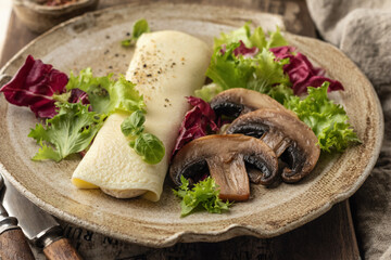 French omelet roll filled with cream cheese with green salad and mushrooms for breakfast on rustic wooden board close up