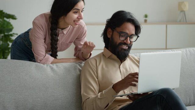 Arabian man businessman freelancer working in home office Indian woman wife stand behind husband multiracial couple family make internet order search apartment on laptop estate website shopping online