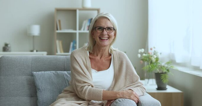 Blonde mature woman in casual clothes smile pose for camera seated on sofa in living room. Happy single senior female relax at own home feel satisfied with carefree retired life and social insurance