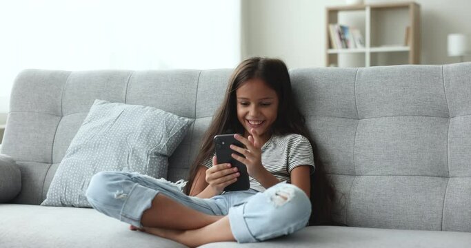 Cheerful little girl relax at home sits on cozy couch in living room, hold smartphone, watch funny video content in social media network, web surfing, chat, use on-line app, enjoy communication. Tech