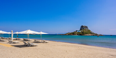 Deck chair and umbrella on beautiful Agios Stefanos Beach in front of paradise Island Kastri- historical ruins and paradise scenery at coast of island Kos, Greece - 611789581