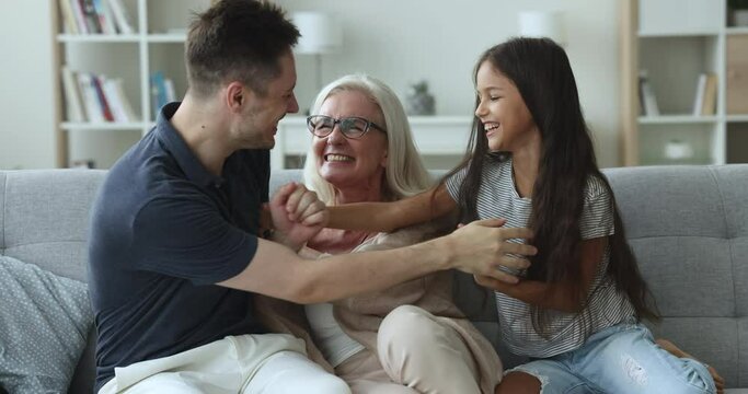Laughing multigenerational family enjoy playtime on couch tickling each other, spend leisure together. Little girl play with daddy and mature granny at home feel happy on weekend, relish communication