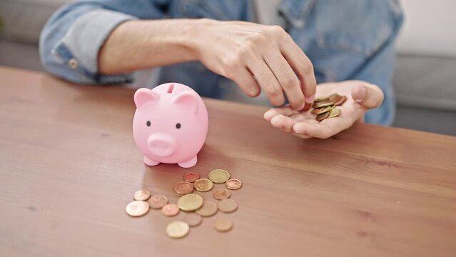 Young hispanic man inserting coins on piggy bank sitting on floor at home