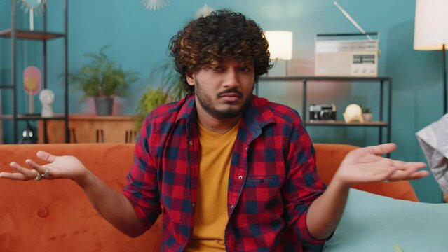 I dont know what to say. Confused indian handsome man feeling embarrassed about ambiguous question, having doubts, no idea, being clueless and uncertain at home. Hindu guy sits on sofa in living room