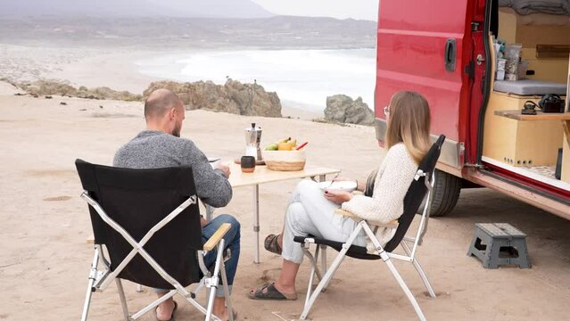 Caucasian adult couple sitting outside at camping table having breakfast next to camper trailer