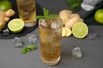 Glass of tasty ginger ale with ice cubes and ingredients on grey wooden table