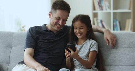 Friendly loving family of dad and cute little child relax sit on sofa in living room, use smartphone for pastime, watch video in social network, have fun with new application, enjoy warm relationships