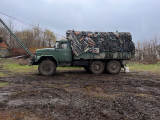 large military truck covered with camouflage net against background of an old fence and dirty soil from rain. War in Ukraine