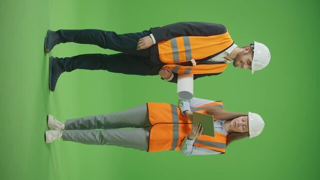 Vertical Footage.Green Screen.Young Smiling Male and Female Civil Engineers in Safety Jackets,White Helmets with Tablet and Blueprints, Discussing Project Work with Interest. Team Work Concept