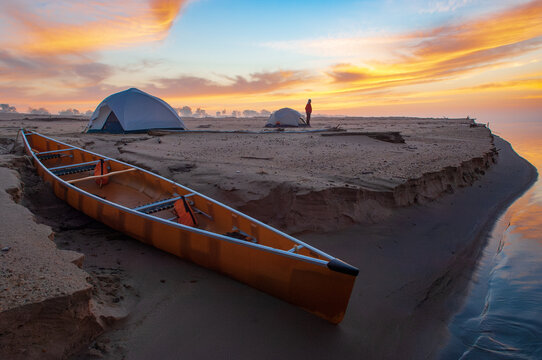 A woman emerges from her tent at sunrise near a canoe on the sandy shore of along the Mississippi River