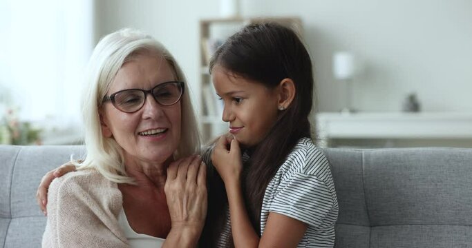 Pretty 10s granddaughter spend weekend time with old attractive granny sitting on couch, girl whisper girlish secrets in ears, secretly talking to grandma, enjoy gossips. Family relationship, trust