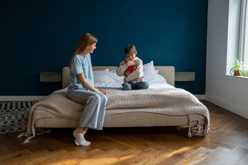 Loving young caucasian mother congratulating happy cute little boy son with wrapped gift. Excited kid sitting on bed getting present, child celebrating birthday with mom at home