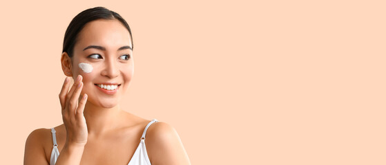 Beautiful Asian woman applying cream on her face against beige background with space for text
