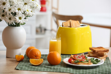 Modern toaster with crispy bread slices, glass of juice, orange and delicious sandwiches on table in kitchen