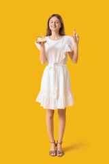 Pretty young woman with birthday cake making wish on yellow background