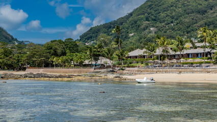Fototapeta na wymiar Low tide on a tropical beach. The rocks of the seabed were exposed. The white boat is moored in shallow water. On the shore the sun umbrellas, the villas of the hotel are visible. Seychelles. Mahe.