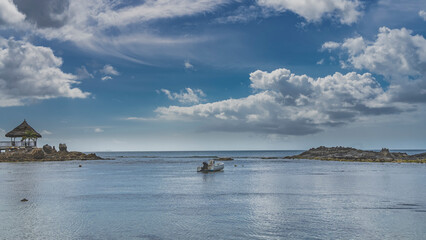 The rocks of the seabed were exposed at low tide. A boat is moored in the lagoon in shallow water. Gazebo with a thatched roof on a background of blue sky and clouds. Seychelles. Mahe.