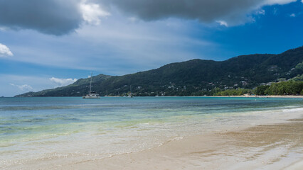 The waves of the turquoise ocean spread over the beach. Wet sand glistens. Yachts in the distance. A green hill against a background of blue sky and clouds. Seychelles. Mahe. Beau Vallon.