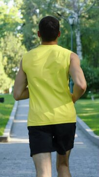 Vertical Screen: Athletic man wearing yellow shirt and black shorts jogging in park in the morning. Following shot jogger exercising outside in summer. Concept of fitness