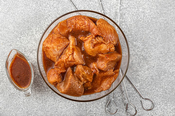 Raw meat marinated meat with bbq sauce in a bowl with skewers for cooking barbecue on gray background