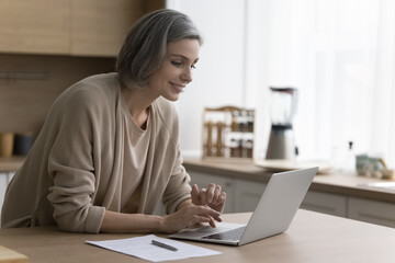 Cheerful mature freelancer woman working from home, using job application on laptop computer, enjoying internet technology for remote freelance job, online communication, smiling