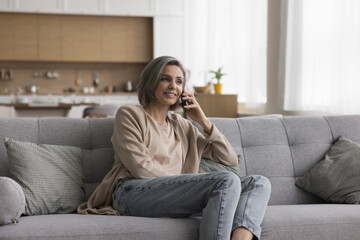 Happy middle aged woman making telephone call from home, talking on mobile phone, enjoying leisure, cellphone conversation, communication, resting on sofa, smiling, laughing