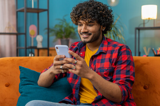 Indian man sitting on sofa uses mobile phone smiles at home living room apartment. Hindu young guy texting share messages content on smartphone social media applications online, watching relax movie