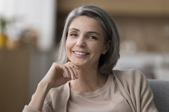 Happy beautiful mature woman indoor head shot portrait. Positive naturally grey haired middle aged touching chin, face, looking at camera with thoughtful toothy smile, resting, sitting on home sofa