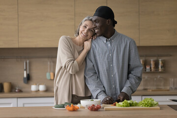 Cheerful cute diverse mature couple keeping healthy lifestyle, preparing salad from fresh vegetables, using organic ingredients, posing at table, looking at camera, smiling, laughing, hugging - Powered by Adobe