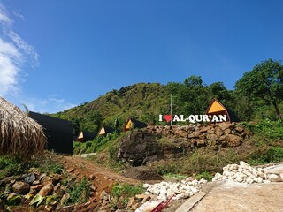 A beautiful photo showcasing a rocky hill with the words "I Love Alquran" written on it, surrounded by lush green trees, a blue sky, and brown soil.