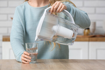 Woman pouring water into glass from filter jug at table in kitchen - obrazy, fototapety, plakaty