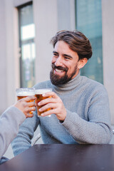 Vertical portrait of a happy young adult man clinking a beer glass with his partner. One cherful caucasian handsomemale drinking and toasting with a friend at bar terrace outside. High quality photo
