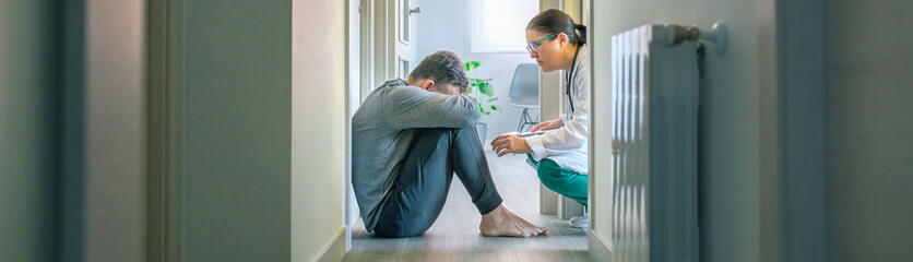 Female psychiatrist talking to male patient with mental disorder and suicidal thoughts sitting on the room floor of hallway - obrazy, fototapety, plakaty