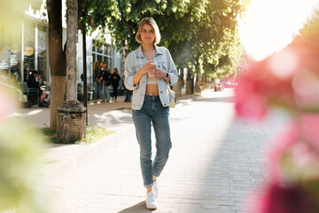 Cute slim young woman in casual clothes with cup of coffee in park on sunny day looking at camera, summer lifestyle