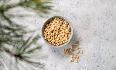 Pine nuts in a bowl and scattered on a light texture background with branches of pine needles . The...
